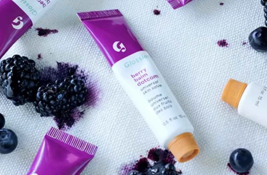 scattered purple and white tubes of Glossier's Balm Dotcom salve with fruit in background