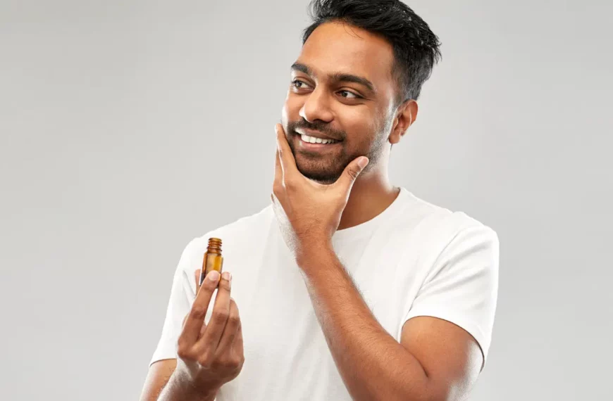 man looking off camera, smiling, touching beard and holding a bottle of beard care product