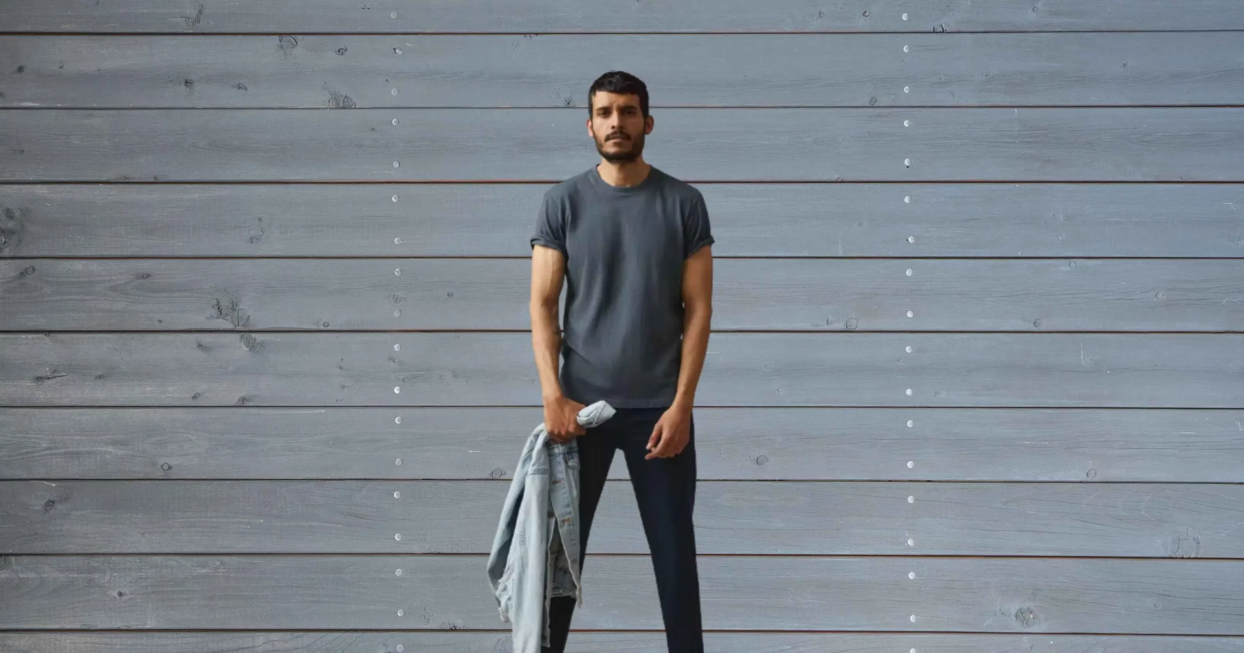 man looking at camera wearing gray shirt, dark pants, holding jean jacket from American Giant