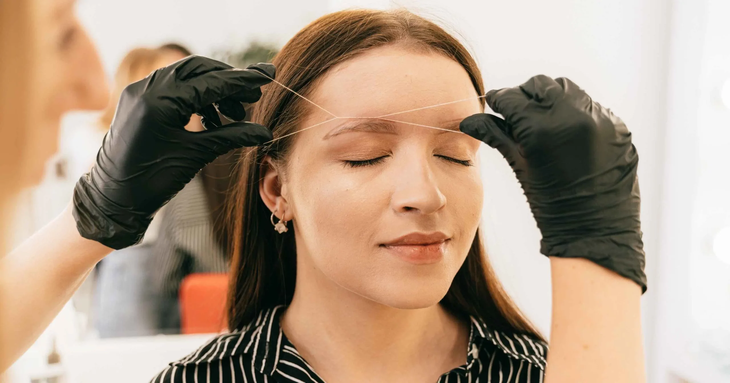 woman with eyes closed having eyebrow threaded by person wearing black gloves