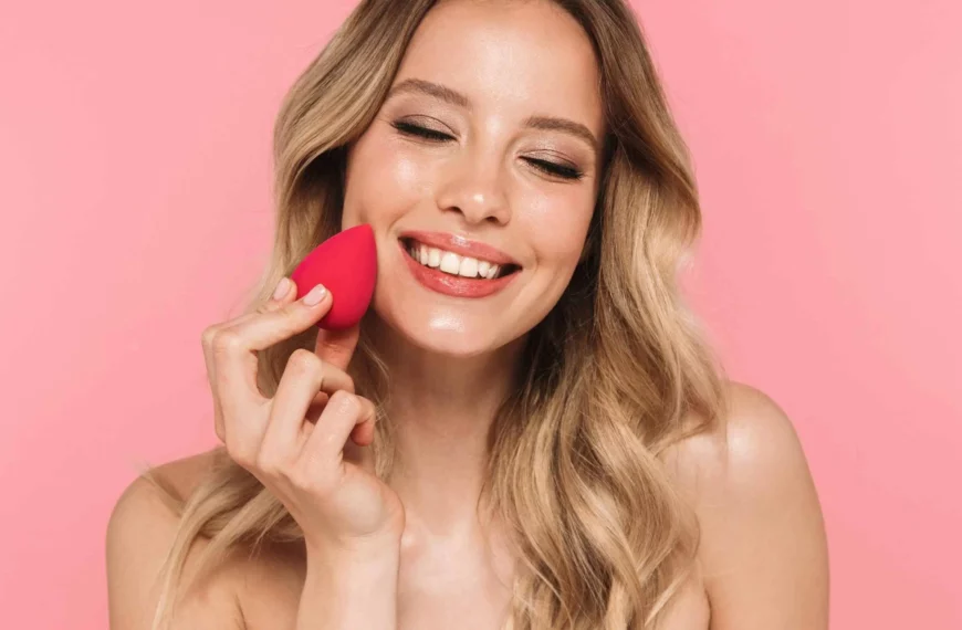 headshot of woman with curly blonde hair, closing eyes, using beauty blender to apply foundation to skin