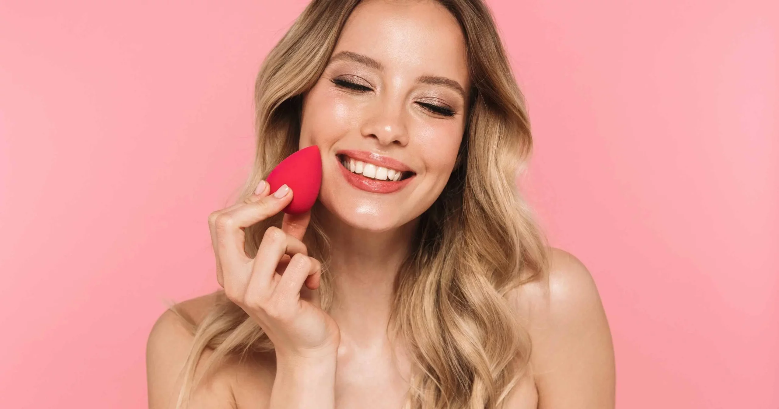 headshot of woman with curly blonde hair, closing eyes, using beauty blender to apply foundation to skin