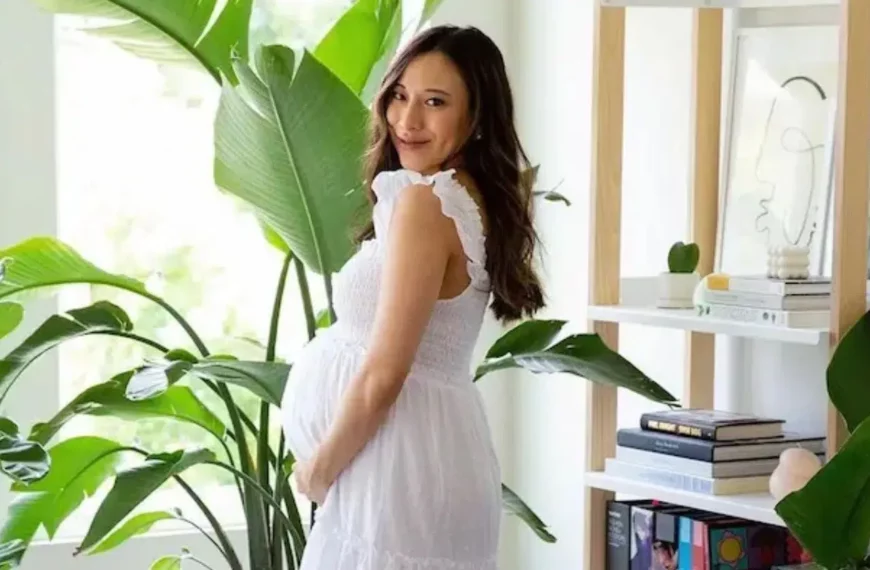 pregnant women looking at camera, holding belly, wearing white maternity clothes dress