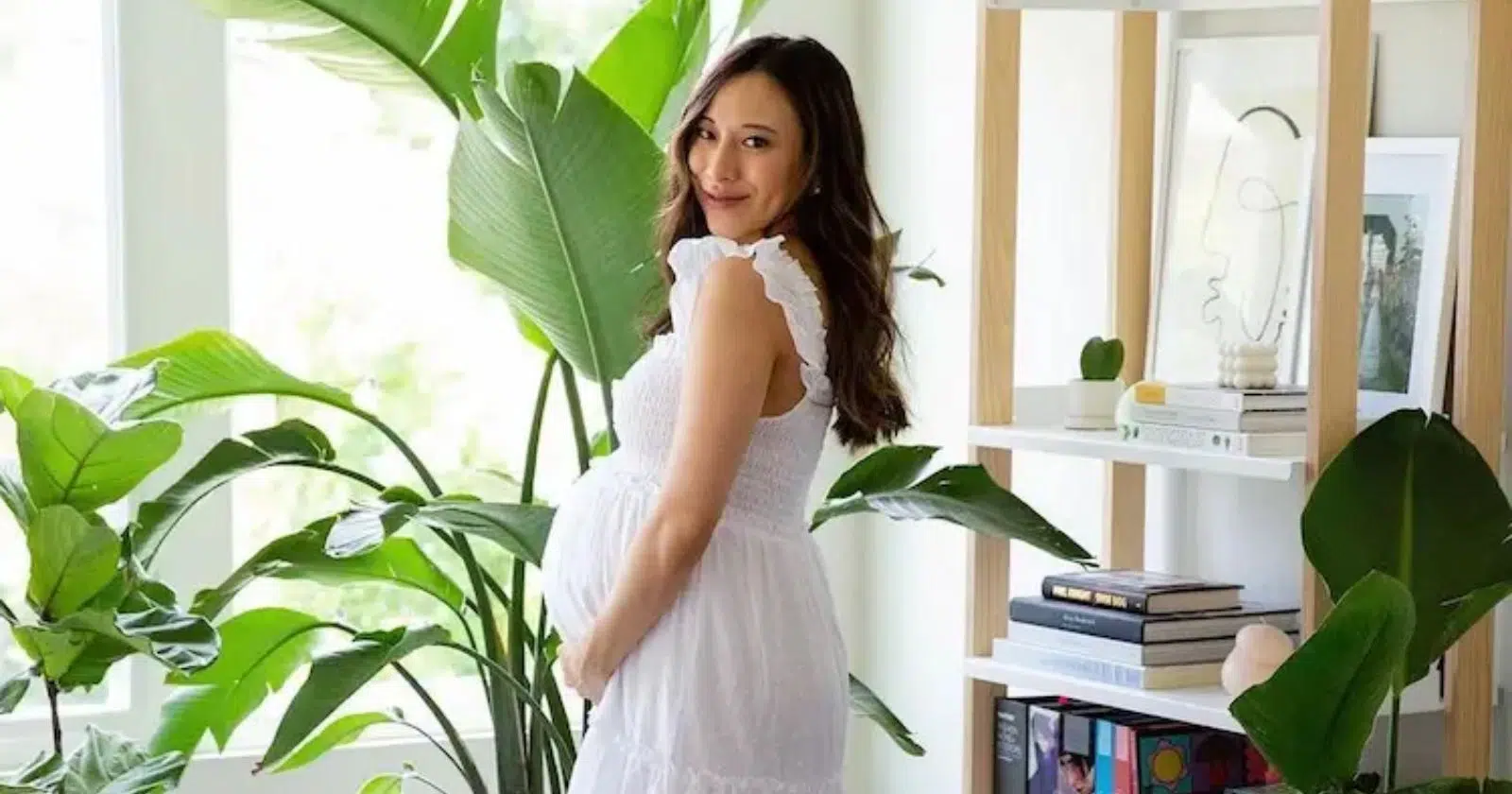 pregnant women looking at camera, holding belly, wearing white maternity clothes dress