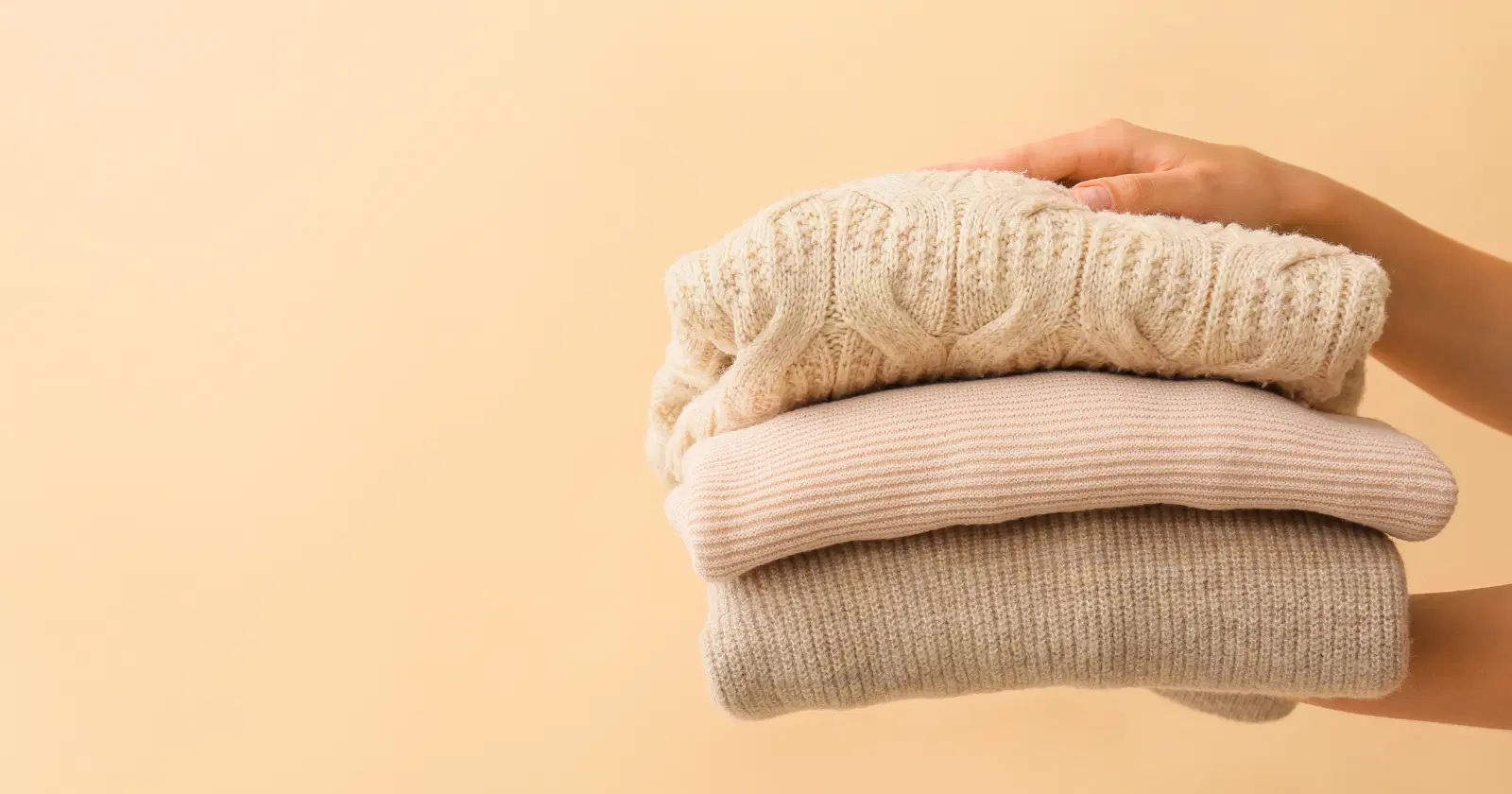 pair of hands holding out a stack of neutral colored polyester sweaters