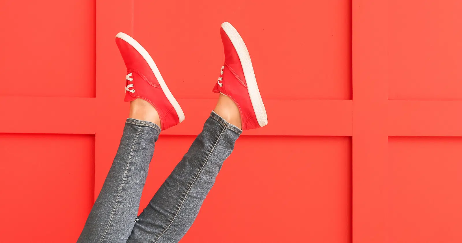 person's feet in the air, wearing red shoes and blue jeggings against an orange-red background